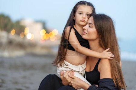 Foto de Serena madre joven abrazando a su pequeña hija con los ojos cerrados, mientras está sentada en la playa. Familia feliz juntos en vacaciones, verano, concepto de viaje - Imagen libre de derechos