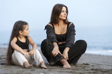Foto de Madre joven y romántica hija pequeña abrazándose, sentada en la playa y mirando hacia otro lado. Familia feliz relajándose de vacaciones - Imagen libre de derechos