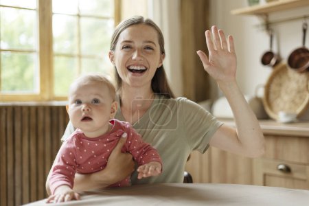 Photo for Smiling young mother holding little cute baby having video call, waving hand, sitting in kitchen, working from home, remote work. Beautiful female watching video. Concept of communication, motherhood - Royalty Free Image