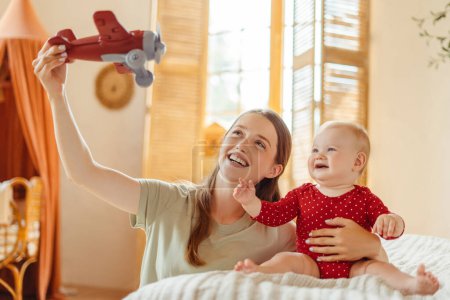 Téléchargez les photos : Jeune baby-sitter heureux jouant avion avec mignon petit bébé assis sur un lit confortable à la maison. Développement du bébé, concept de parentalité - en image libre de droit