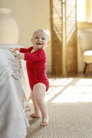 Photo for Portrait of cute little toddler wearing red baby clothes standing alone near bed at cozy light home. Firs step concept - Royalty Free Image