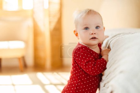 Téléchargez les photos : Portrait de mignon petit tout-petit portant des vêtements de bébé rouge debout près du lit à la maison. Concept de la première étape - en image libre de droit