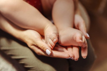 Téléchargez les photos : Vue recadrée de la jeune mère tenant ses jolies petites jambes de bébé tout en étant assise à la maison. Amour, soins, concept de maternité - en image libre de droit