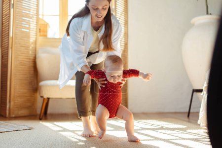Photo for Young smiling mother teaching to walk her toddler at home. Happy family together. First step, baby development concept - Royalty Free Image