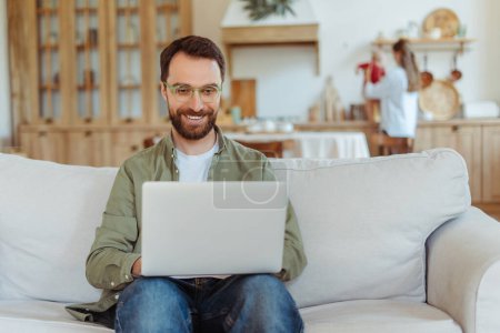 Téléchargez les photos : Portrait d'homme barbu souriant, pigiste portant des lunettes élégantes à l'aide d'un ordinateur portable travaillant en ligne depuis la maison. Technologie, concept d'emploi à distance - en image libre de droit