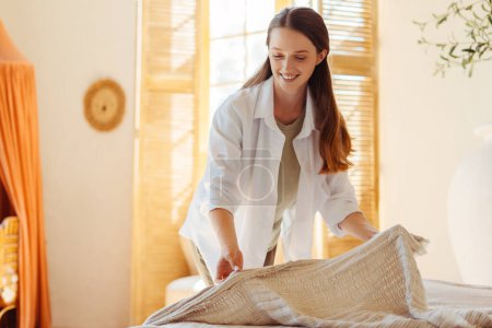 Photo for Beautiful, smiling woman making bed, servant cleaning bedroom. Attractive female wearing casual clothes. Cute saleswoman presenting blanket. Concept of service, shopping - Royalty Free Image