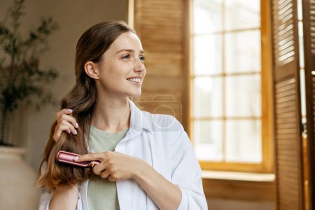 Photo for Young smiling woman combing her beautiful long hair hair looking away at home. Hair care, morning routine concept - Royalty Free Image