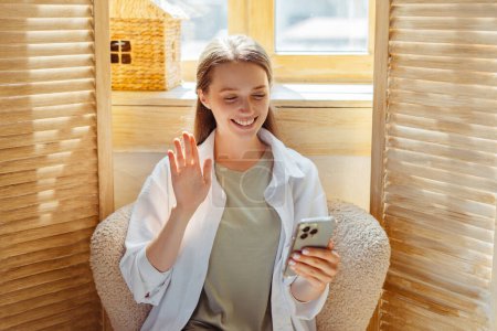 Foto de Retrato sonriente mujer sosteniendo la comunicación del teléfono móvil en línea, saludando la mano, teniendo videollamada sentado en el sillón en casa. Influencer grabación de vídeo con smartphone. Redes sociales - Imagen libre de derechos