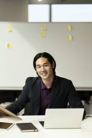 Téléchargez les photos : Bel homme asiatique souriant à l'aide d'un ordinateur portable regardant la caméra de travail en ligne dans le bureau moderne avec des notes collantes sur fond. Scrum, agile - en image libre de droit