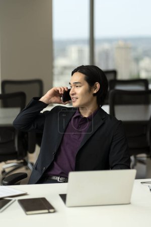 Téléchargez les photos : Portrait d'un bel homme d'affaires asiatique souriant, gestionnaire parlant sur un téléphone mobile, répondant à un appel assis dans un bureau moderne. Concept technologique - en image libre de droit