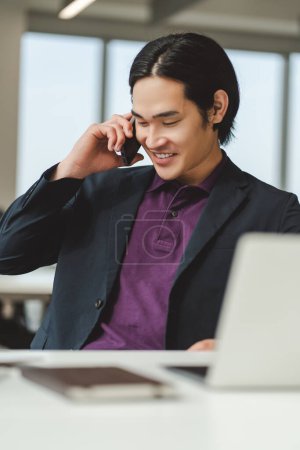 Foto de Retrato de un exitoso joven empresario asiático usando un portátil, hablando por teléfono móvil trabajando en una oficina moderna. Elegante freelancer sentado en el lugar de trabajo. Concepto de comunicación, tecnología - Imagen libre de derechos