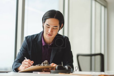 Foto de Retrato del hombre de negocios asiático tomando notas sentado en el lugar de trabajo en la oficina moderna. Estudiante japonés atractivo estudiando, proyecto de planificación, trabajando desde casa. Concepto educativo - Imagen libre de derechos
