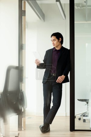 Foto de Guapo seria asiático hombre sosteniendo digital tablet trabajo en línea de pie en moderno oficina. Concepto tecnológico - Imagen libre de derechos