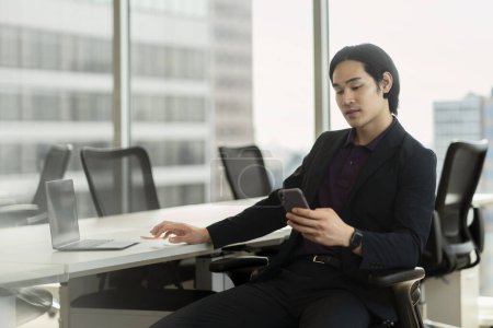 Foto de Guapo pensativo asiático hombre holding móvil lectura texto mensaje sentado en moderno oficina - Imagen libre de derechos