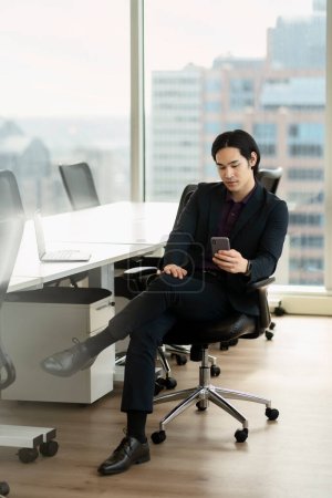 Foto de Retrato de un empresario asiático serio y seguro que sostiene el teléfono móvil trabajando en línea, leyendo el mensaje de texto, verificando el correo electrónico sentado en la silla de la oficina. Tecnología, concepto de negocio exitoso - Imagen libre de derechos