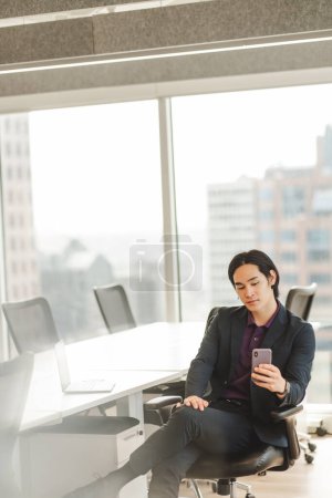 Foto de Retrato de un serio hombre de negocios asiático sentado en el escritorio, sosteniendo el teléfono móvil, navegando en la oficina moderna. Atractivas compras en línea masculina, comercio electrónico. Concepto tecnológico - Imagen libre de derechos