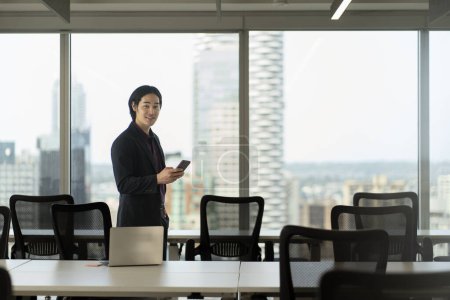 Photo for Portrait of handsome smiling asian man holding smartphone using mobile app standing in modern office. Technology, successful business concept - Royalty Free Image