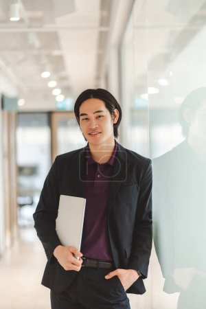 Téléchargez les photos : Portrait de beau gestionnaire homme asiatique portant costume noir élégant tenue formelle ordinateur portable, en regardant la caméra. Modèle masculin japonais attrayant posant dans un bureau moderne. Concept de technologie d'entreprise - en image libre de droit
