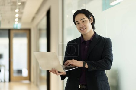 Foto de Sonriente hombre asiático con traje negro sosteniendo ordenador portátil de pie en la oficina. Tecnología, concepto de negocio exitoso - Imagen libre de derechos