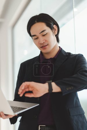 Téléchargez les photos : Portrait de succès asiatique homme d'affaires en utilisant ordinateur portable de travail en ligne, projet de planification, vérification du temps d'attente dans le bureau moderne. Homme d'affaires intelligent sur le lieu de travail, concept d'entreprise technologique - en image libre de droit