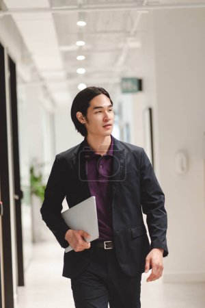 Foto de Guapo, confiado hombre de negocios asiático, director ejecutivo en traje negro elegante que sostiene el ordenador portátil, caminando por la oficina mirando hacia otro lado. Atractivo gerente financiero japonés. Concepto empresarial - Imagen libre de derechos
