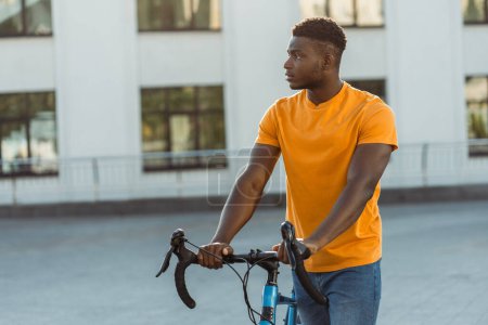Photo for Dark skinned handsome man walking street near bicycle while looking away. Calm african american student crossing with bike - Royalty Free Image