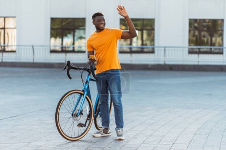 Photo for Porter of smiling positive Nigerian man waving hand standing near bicycle holding mobile phone. Handsome male wearing stylish yellow t shirt on urban street. Concept of active healthy lifestyle - Royalty Free Image