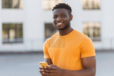 Photo for Portrait of handsome african man holding mobile phone, looking at camera outdoors, on urban street. Successful african american businessman looking at camera. Concept of online communication - Royalty Free Image