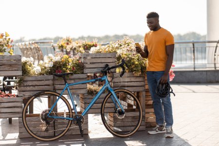 Foto de Retrato de un hombre afroamericano guapo sonriente sosteniendo un teléfono móvil, tomando fotos de pie cerca de la bicicleta en la calle. Atractivo chico africano sosteniendo casco de seguridad, compras en línea al aire libre, ciclismo - Imagen libre de derechos