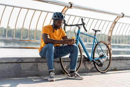 Foto de Feliz hombre multirracial con casco protector sentado en la calle y sosteniendo el teléfono móvil mientras se relaja después de montar en bicicleta - Imagen libre de derechos