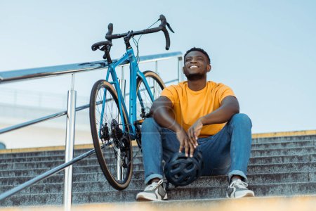 Téléchargez les photos : Portrait d'un Africain positif souriant se reposant, assis sur des escaliers, bicyclette près de lui. Beau hipster heureux en vêtements décontractés à l'extérieur. Concept de mode de vie sain actif - en image libre de droit