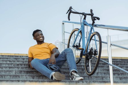 Téléchargez les photos : Un Afro-Américain souriant portant des vêtements orange élégants couché, assis sur des escaliers avec un vélo à côté de lui. Attrayant mâle tenant casque se reposant après le vélo. Concept de mode de vie sain - en image libre de droit
