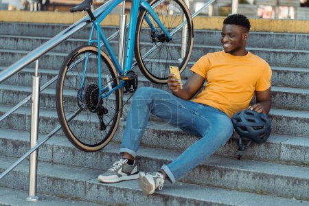 Photo for Happy african american man looking at mobile phone screen while relaxing at the stairs after bicycle riding. People lifestyle concept - Royalty Free Image