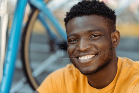 Photo for Portrait of attractive smiling Nigerian male rider, wearing orange t shirt, copy space. Positive young student sitting near bicycle, closeup. Handsome male posing for picture. Cycling concept - Royalty Free Image