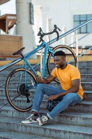 Téléchargez les photos : Attrayant souriant homme afro-américain assis sur des escaliers près de vélo, tenant téléphone mobile, achats en ligne, à l'extérieur. Jeune cavalier en utilisant smartphone, détente, vérifier email. Concept technologique - en image libre de droit