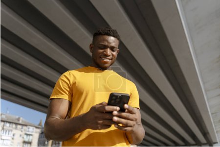 Téléchargez les photos : Un jeune homme afro-américain positif tenant un téléphone portable, regardant une vidéo dans la rue de la ville. Charmant homme d'affaires africain souriant vérifiant le courrier. Concept de communication en ligne - en image libre de droit