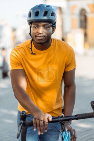 Photo for Portrait of smiling African rider wearing protective helmet, walking with bicycle on urban street, closeup. Attractive hipster in casual yellow t shirt looking away. Cycling concept, healthy lifestyle - Royalty Free Image