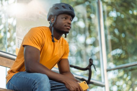 Téléchargez les photos : Souriant homme afro-américain regardant loin tout en se relaxant à vélo à la gare routière après avoir roulé. Concept de style de vie - en image libre de droit
