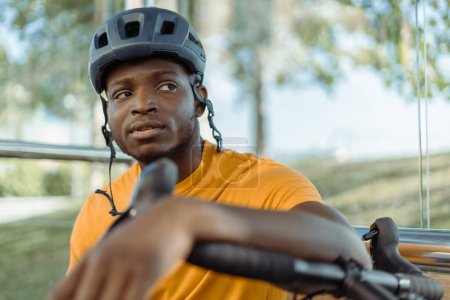 Photo for Portrait of handsome African American man wearing safety helmet sitting at bus stop, looking away. Pensive Nigerian tourist thinking about travel. Transportation concept - Royalty Free Image