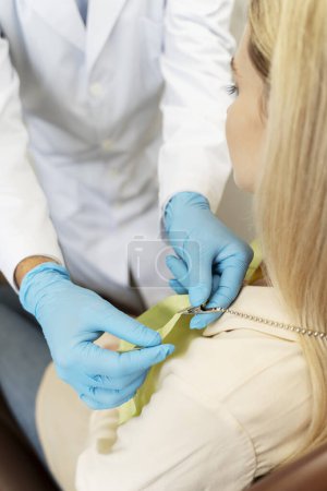 Photo for Dentist attaching dental napkin to attractive female patient with blond hair in modern clinic. Concept of dental care, health care - Royalty Free Image