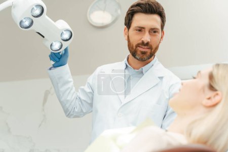 Téléchargez les photos : Dentiste barbu professionnel tournant la lampe tout en préparant vérifier les dents de la femme. Jeune belle patiente souriante assise dans une clinique dentaire moderne. Traitement des dents, concept de soins de santé - en image libre de droit