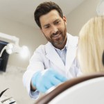 Handsome bearded dentist preparing female patient to checking teeth. Back view of the blonde woman sitting in modern dental clinic. Teeth treatment, health care concept 