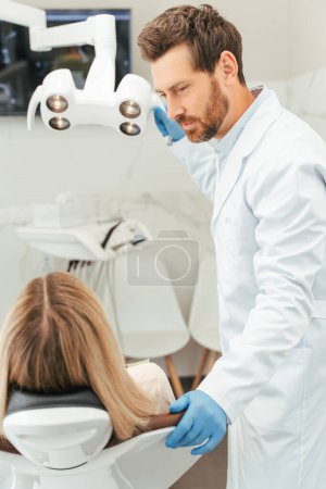 Téléchargez les photos : Portrait d'un dentiste séduisant d'âge moyen consultant une patiente aux cheveux blonds, assise dans une chaise dentaire. Médecin professionnel préparant lampe dentaire. Concept de traitement, soins dentaires - en image libre de droit
