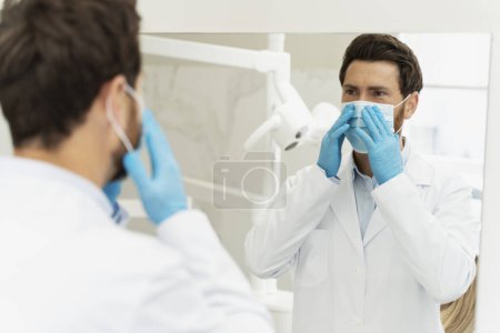 Photo for Confident dental practitioner, dentist, stomatologist orthodontist, wearing medical mask and uniform, looking at mirror while preparing for regular check up - Royalty Free Image