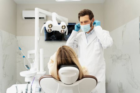 Téléchargez les photos : Dentiste professionnel portant un masque protecteur tout en se préparant au contrôle des dents. Patiente blonde assise dans une clinique dentaire moderne. Traitement des dents, concept de soins de santé - en image libre de droit