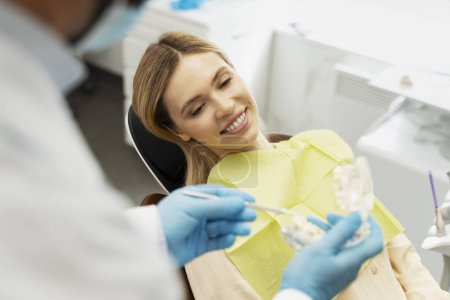 Téléchargez les photos : Portrait d'une belle patiente séduisante avec un sourire de dents lors d'une visite chez le dentiste, assise dans une chaise dentaire. Médecin montrant la mâchoire en plastique, traitement. Concept de soins dentaires - en image libre de droit