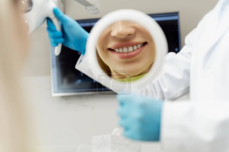 Foto de Retrato de paciente femenina con sonrisa dentada mirando en espejo después del tratamiento dental. Mujer positiva visitando dentista. Cuidado dental, concepto de higiene bucal - Imagen libre de derechos