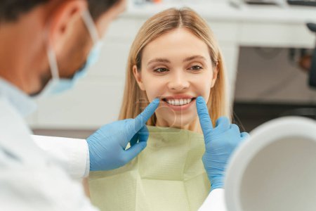 Téléchargez les photos : Portrait d'une belle femme caucasienne souriante regardant dans un miroir, vérifiant ses dents dans une clinique dentaire moderne. Concept de soins de santé - en image libre de droit
