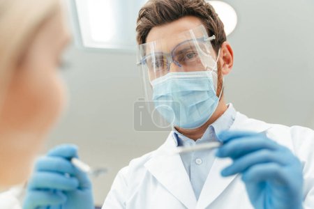 Téléchargez les photos : Portrait d'un beau dentiste masculin d'âge moyen en masque protecteur et lunettes, gants en latex vérifiant les dents de la patiente. Traitement, soins de santé, dentisterie concept - en image libre de droit