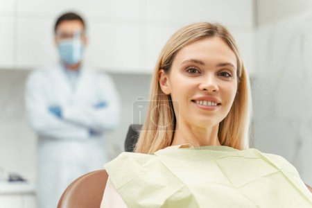 Foto de Retrato de una hermosa mujer rubia sonriente sentada en una clínica dental moderna con dentista en el fondo. Tratamiento dental, concepto de cuidado de la salud - Imagen libre de derechos